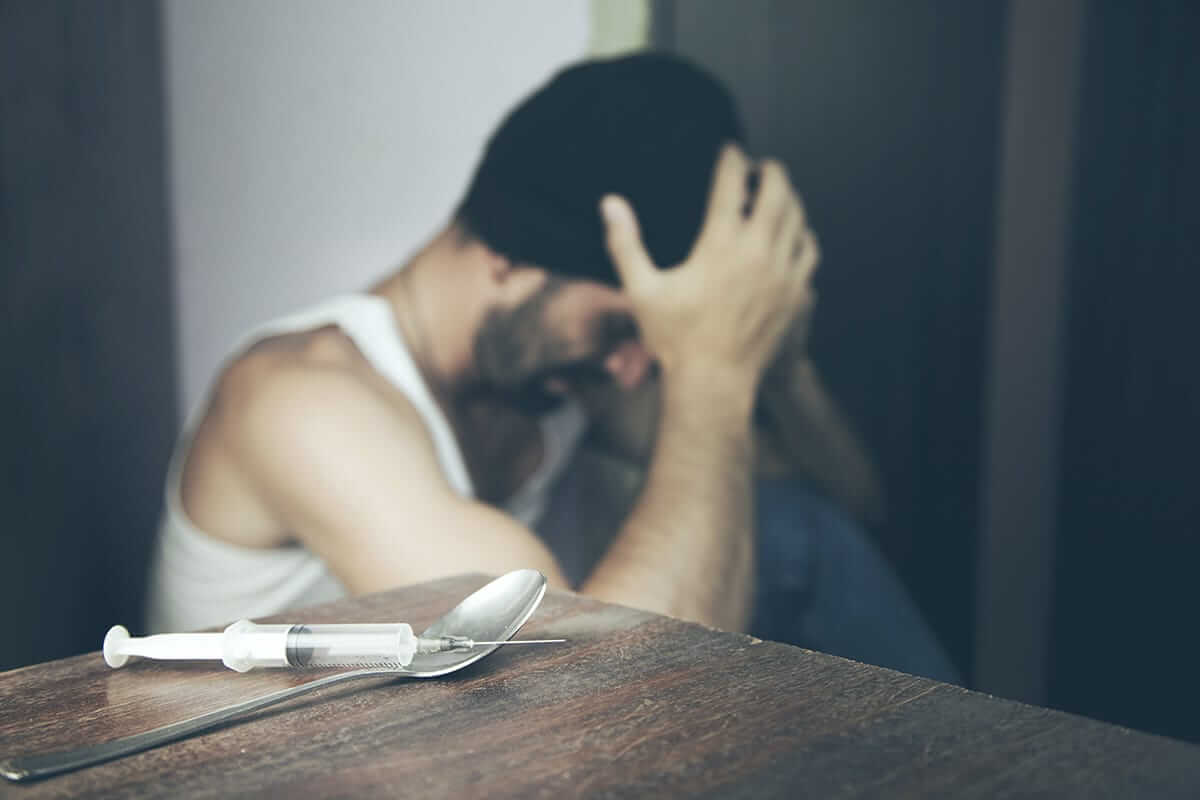 man in beanie sits with hands on his head with syringe and spoon on table debating if it's heroin vs fentanyl